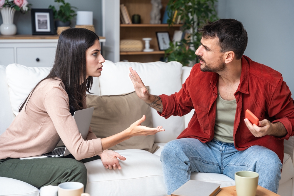 A man and a woman are engaged in a heated conversation while sitting on a couch in a cozy living room. The woman is expressing frustration with her hands raised, while the man is gesturing with an outstretched hand, appearing defensive.